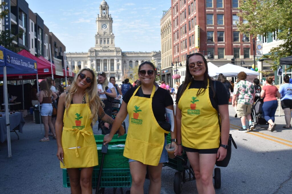 three volunteers pose with a wagon full of produce donations in front of the court house and farmers market booths.