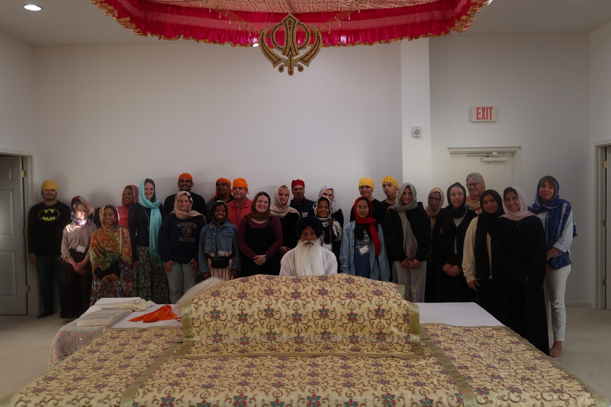 A group of about 20 students stand with Sikhi men in front of the holy word in the Gurdwara